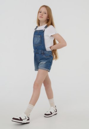 Niebieskie ogrodniczki dżinsowe z regulowanymi ramiączkami, kieszenią z przodu i podwijanymi nogawkami. W zestawie z białą koszulką i sportowymi butami.