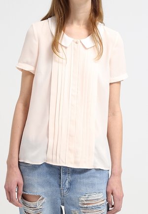 Blouse rose clair avec un col blanc, dotée de plis verticaux et de manches courtes, associée à un jean en denim bleu déchiré.