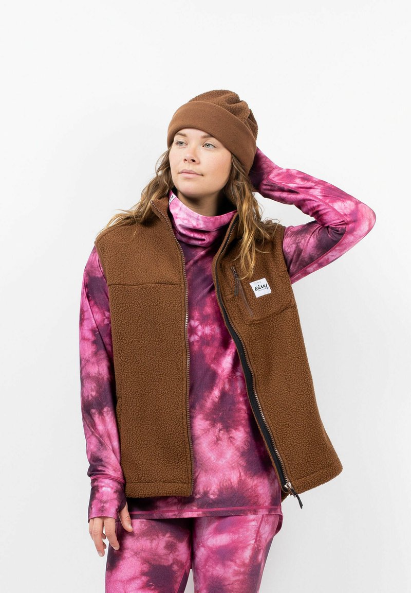 Bruine fleece vest met ritszak, gecombineerd met een roze tie-dye lange mouw shirt en bijpassende leggings. Bruine beanie op het hoofd.