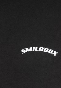 Bílé vyšívané „SMILODOX“ text na černé texturované tkanině, umístěné uprostřed a mírně zakřivené směrem nahoru.