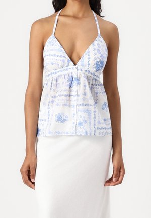 Femme portant un top halter blanc avec des motifs floraux bleus, assorti à une jupe blanche unie sur un fond uni.