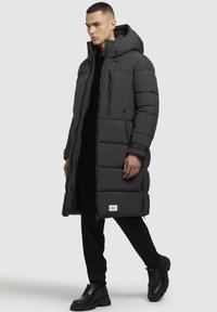 Lang sort pufferjakke med hætte, med lynlås fortil, horisontalt quiltet design og sidelommer, iført over sorte bukser og støvler.