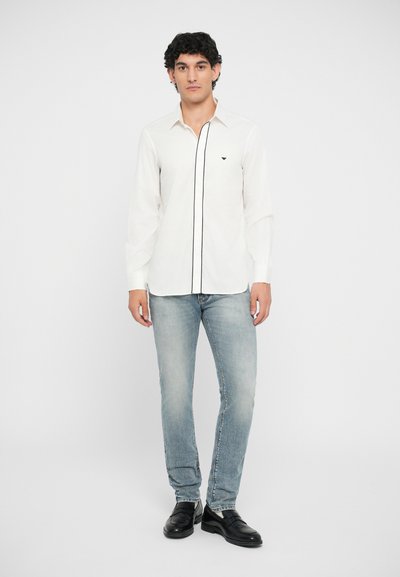 Vit långärmad skjorta med svart kant och liten logotyp, kombinerad med ljusblå jeans och svarta skor. Modellen står mot en enfärgad bakgrund.