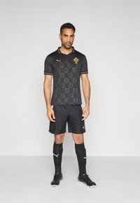 Camisola de futebol preta com logótipo dourado, design com padrão e detalhes verde-vermelho. Combinada com calções pretos, meias e chuteiras pretas.