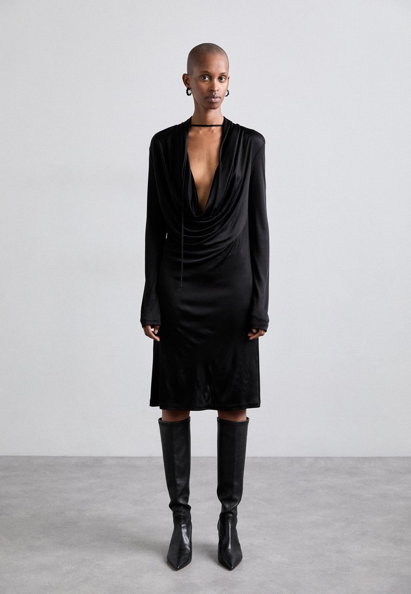 Rochie neagră, drapată, cu decolteu adânc în V și mâneci lungi. Textură netedă, design până la genunchi, asortată cu cizme negre din piele înalte.