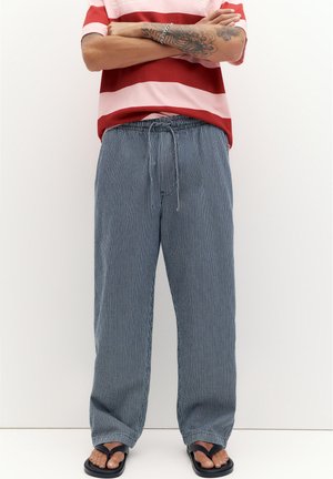 Person iført løse blåternede bukser med snøre, en rød og pink stribet skjorte, sorte sandaler og et sølvarmbånd, stående med armene over kors.