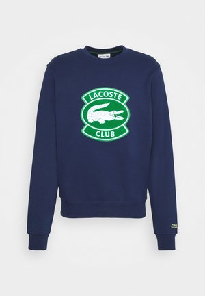 Sweatshirt bleu marine à manches longues, arborant un grand logo Lacoste Club vert et blanc avec un alligator sur la poitrine et un petit alligator sur le poignet.