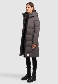 Dark grey padded long coat with a hood, featuring a front zipper and snap buttons, and side zip pockets. Black knee-high boots complete the look.