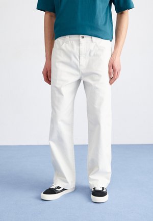 Witte katoenen jeans met een relaxte fit, vijf zakken en een knoopsluiting, gecombineerd met zwart-witte sneakers op een blauwe achtergrond.