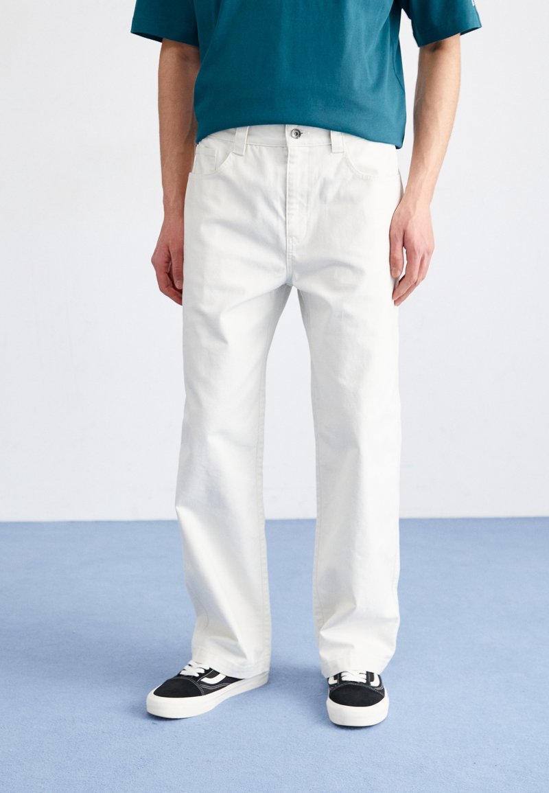 Vaqueros de algodón blancos con un corte relajado, cinco bolsillos y cierre de botón, combinados con zapatillas en blanco y negro sobre un fondo azul.