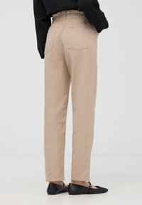 Pantaloni beige a vita alta con vita arricciata, due tasche posteriori e gambe affusolate, abbinati a scarpe nere flat.