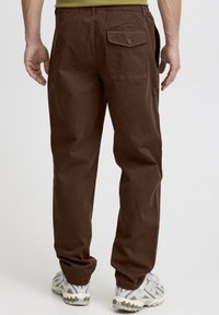 Pantaloni di cotone marroni con una vestibilità ampia, dotati di una tasca posteriore con chiusura a bottone e gambe leggermente aderenti. Indossati con sneaker grigio-bianco.