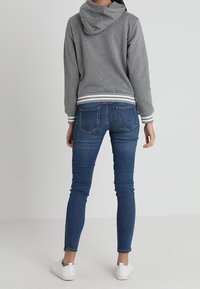 Sudadera gris con capucha y puños a rayas blancas, combinada con unos jeans azules ajustados y zapatillas blancas, vista desde atrás.