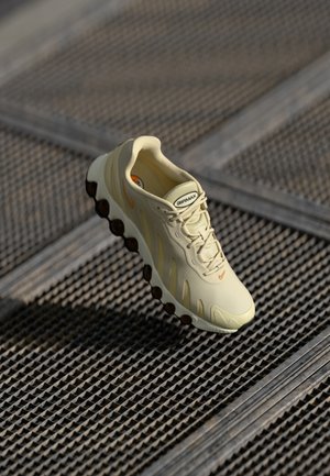 Lysegule sneakers med struktureret overdel, fremtrædende snøring, orange detaljer og en sort boblesål på en gitterbaggrund.