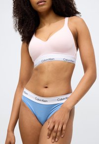 Ροζ ασύρματο σουτιέν με ευρεία λευκή ταινία Calvin Klein· συνδυασμένο με ανοιχτό μπλε μαγιό κάτω μέρος με την ίδια ταινία. Λείο υφασμάτινο υλικό.