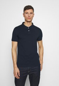 Marineblauwe polo met korte mouwen, met een kraag, twee-knops sluiting en textuurstof. Gedirigeerd met donkere jeans.