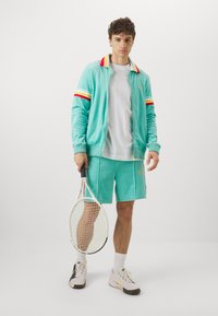 Ensemble de survêtement en velours turquoise avec un col et des poignets rayés multicolores, associé à un short assorti. Tenant une raquette de tennis, portant des baskets légères.