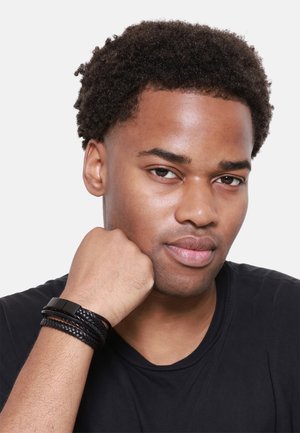 Black leather wrist bracelet featuring braided and smooth textures, worn on a wrist against a dark shirt. Simple, modern design.