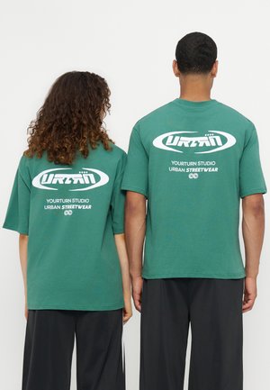 Deux personnes portent des T-shirts verts Yourturn Studio urban streetwear montrant les logos au dos, debout côte à côte sur un fond uni.