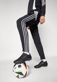 Black soccer cleats on a white sock, fitted black pants with gray stripes, foot resting on a multicolored soccer ball with logos.