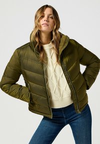Giacca a vento verde oliva con zip frontale, colletto alto e design trapuntato. Indossata sopra un maglione beige a trecce, abbinata a jeans blu.