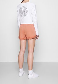 Kort t-shirt med lång ärm i vitt, med en cirkulär logotyp på baksidan, parat med persikofärgade shorts med uppvikta ben och vita sneakers.