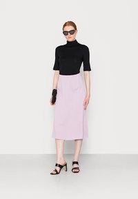 Top preto justo com gola alta, saia midi rosa claro com faixa elástica na cintura e sandálias de salto pretas. Modelo a segurar uma carteira preta.