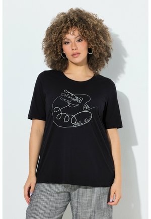 Frau mit lockigem Haar trägt ein schwarzes T-Shirt mit abstrakter weißer Linienzeichnung einer Hand und graue gestreifte Hose, sie steht vor einer weißen Wand.