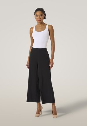 Pantaloni neri a gamba larga con una texture liscia, vita alta e una leggera lucentezza, abbinati a un top bianco aderente e tacchi nudi.