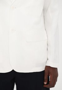 Weiße Leinenblazer mit zwei Knöpfen, einer Brusttasche und taillierter Passform. Der Stoff ist leicht und hat eine glatte Textur.