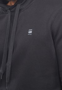 Svart hoodie tillverkad av mjukt tyg, med en ficka framtill, dragsko och en liten logotyp på bröstet. Synlig sömdetaljering.