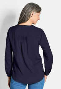 Marinebluse mit langen Ärmeln, die ein rückseitiges Paspel-Detail und einen abgerundeten Saum aufweist. Der glatte Stoff und die geknöpften Manschetten verleihen dem Design eine besondere Note.