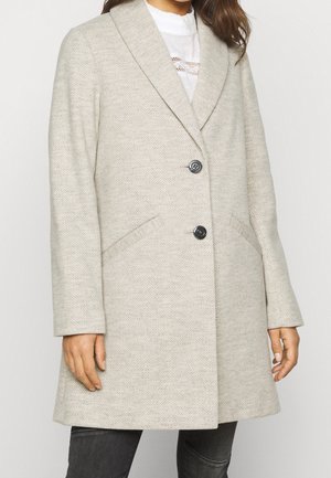 Manteau en laine gris clair à chevrons avec deux boutons noirs, col à revers cranté et poches avant inclinées porté sur un haut blanc et un jean foncé.