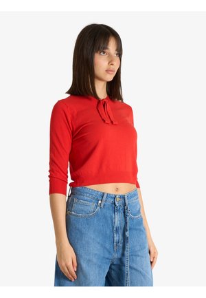 Giovane donna con capelli lisci e scuri che indossa un crop top rosso a maniche lunghe e jeans blu a vita alta su sfondo bianco.