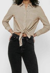 Femme portant une chemise à rayures beige boutonnée et nouée à la taille, avec un jean noir taille haute, debout les mains sur les hanches.