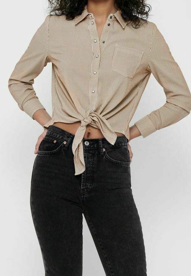 Femme portant une chemise à rayures beige boutonnée et nouée à la taille, avec un jean noir taille haute, debout les mains sur les hanches.