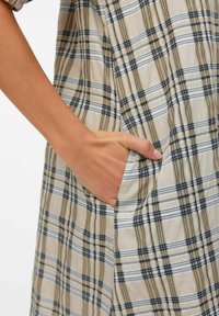 Ręka oparta w bocznej kieszeni beżowej, czarno-białej sukienki w kratkę.