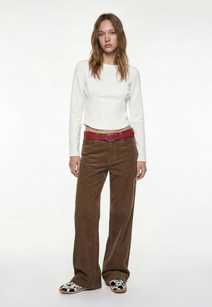 Młoda kobieta ubrana w biały dopasowany top z długim rękawem, brązowe szerokie spodnie z czerwonym paskiem oraz czarno-białe wsuwane buty w cętki.