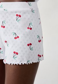 Witte shorts met een rood kersprint en groene bladeren, gemaakt van een gestructureerde stof en met een golvende zoom.