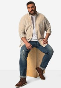 Chemise boutonnée rayée beige sur un t-shirt blanc, associée à un jean bleu déchiré et des bottes à lacets marron, assise sur un piédestal en bois.