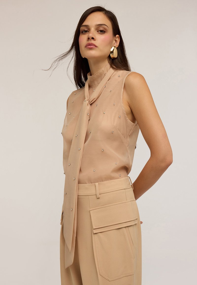Blusa beige senza maniche con collo annodato, adornata con piccoli dettagli in argento; abbinata a pantaloni beige a vita alta con grandi tasche.