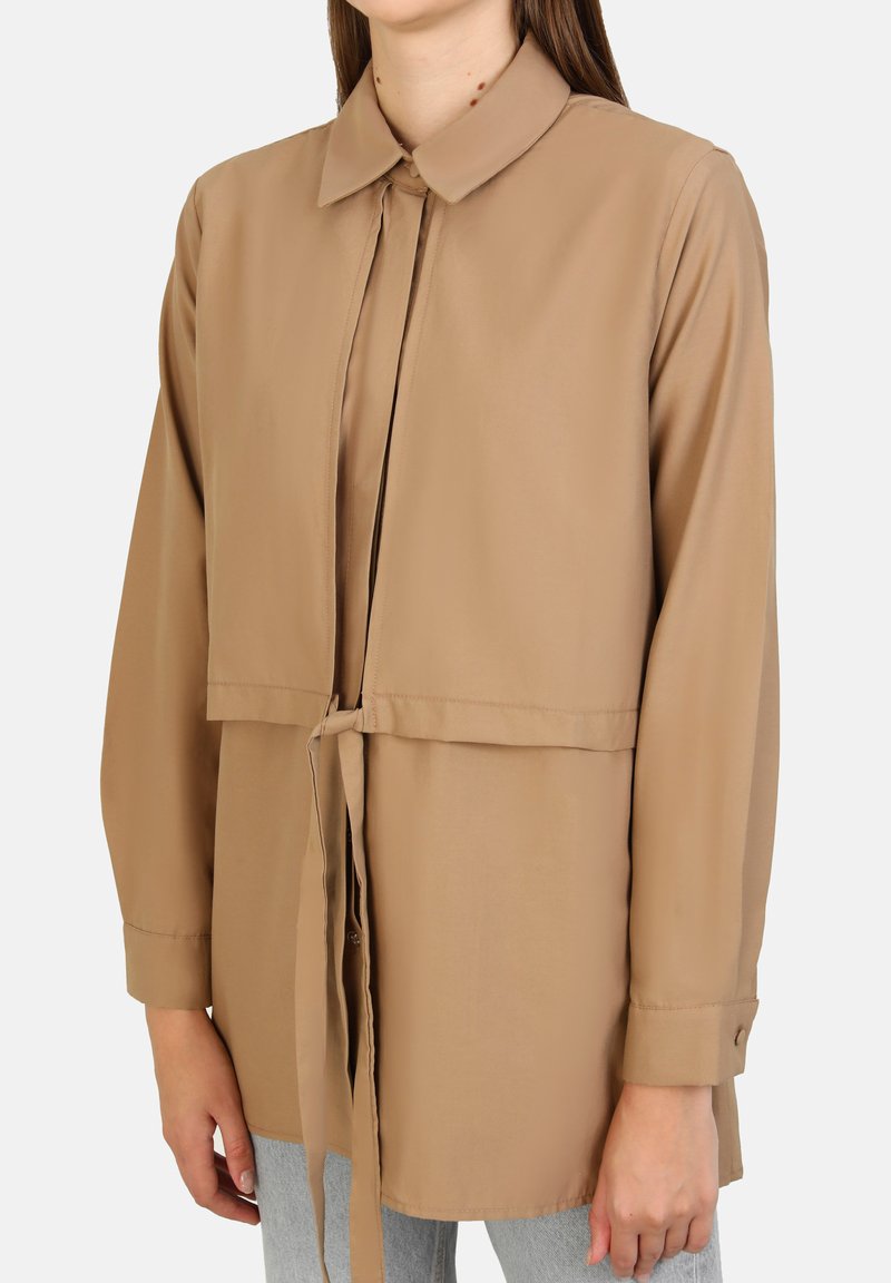 Lichtbruine blouse met een kraag, een strik aan de voorkant en lange mouwen. Gemaakt van gladde stof met een getailleerde pasvorm en een iets verkorte ontwerp.