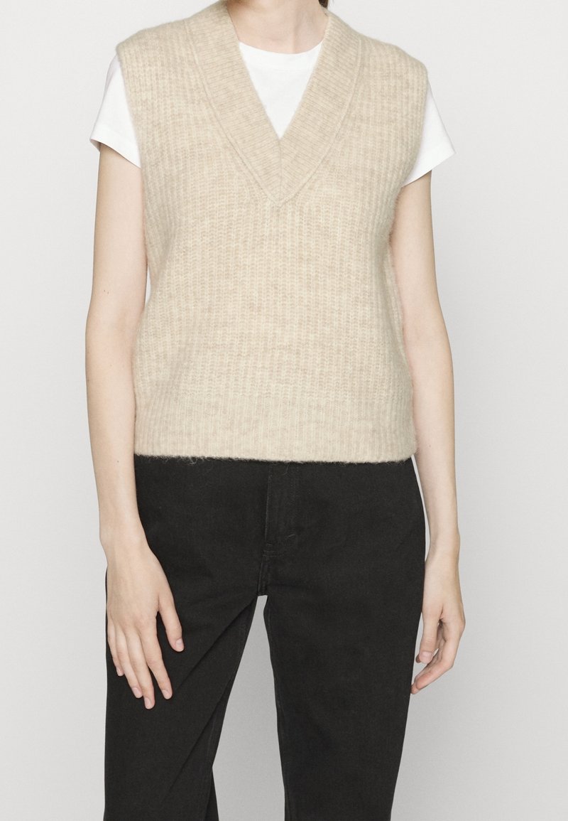 Personne portant un gilet sans manches en maille beige sur une chemise blanche à manches courtes et un pantalon noir, debout devant un fond uni.