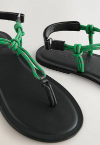 Zwarte platte sandalen met gevlochten groene en roze touwen als bandjes. Voorzien van een teenbeugel en een enkelriem met metalen gespen. Glad, mat oppervlak.