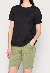 Blusa nera a maniche corte con sottili righe verticali, scollatura rotonda e dettagli arricciati sul seno, abbinata a pantaloni corti verde oliva.