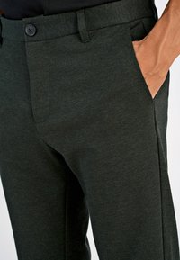 Mørkegrønne bukser med et teksturert stoff, med knappelukking og to sidelommer. Slim fit design som fremhever den skreddersydde formen.