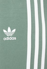 Zielony sportowy top wykonany z miękkiego materiału, z dużym białym logo Adidas oraz trzema białymi pionowymi paskami po lewej stronie.