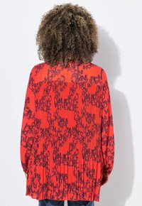 Blusa rossa con motivo di testo nero, schiena plissettata, maniche lunghe e orlo tondo. Tessuto leggero con superficie liscia.