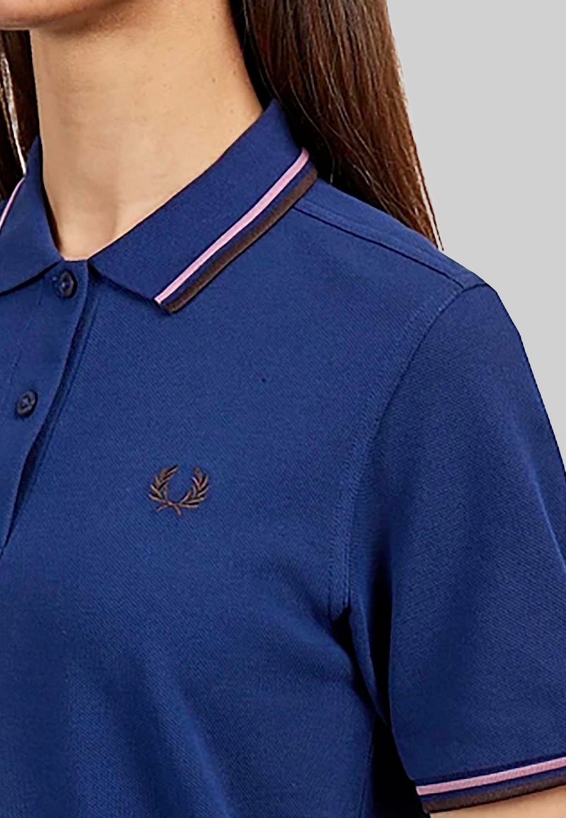 Polo navy avec un col contrastant comportant des rayures roses et brunes. Comprend un petit logo brodé marron sur la poitrine.