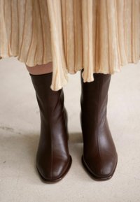 Brown leather ankle boots with a square toe, smooth texture, and vertical stitching, paired with a pleated, light-colored dress.